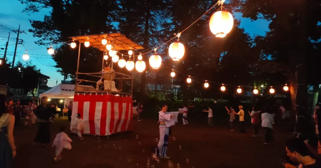 櫓で太鼓を叩く男性たちと、その周りで盆踊りを楽しむ人々が笑顔で踊るどんぐり公園の夏祭り風景